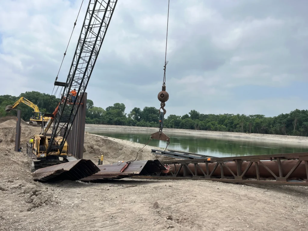 Drainage construction site with a crane, excavator, and steel sheet piles being installed along a canal.