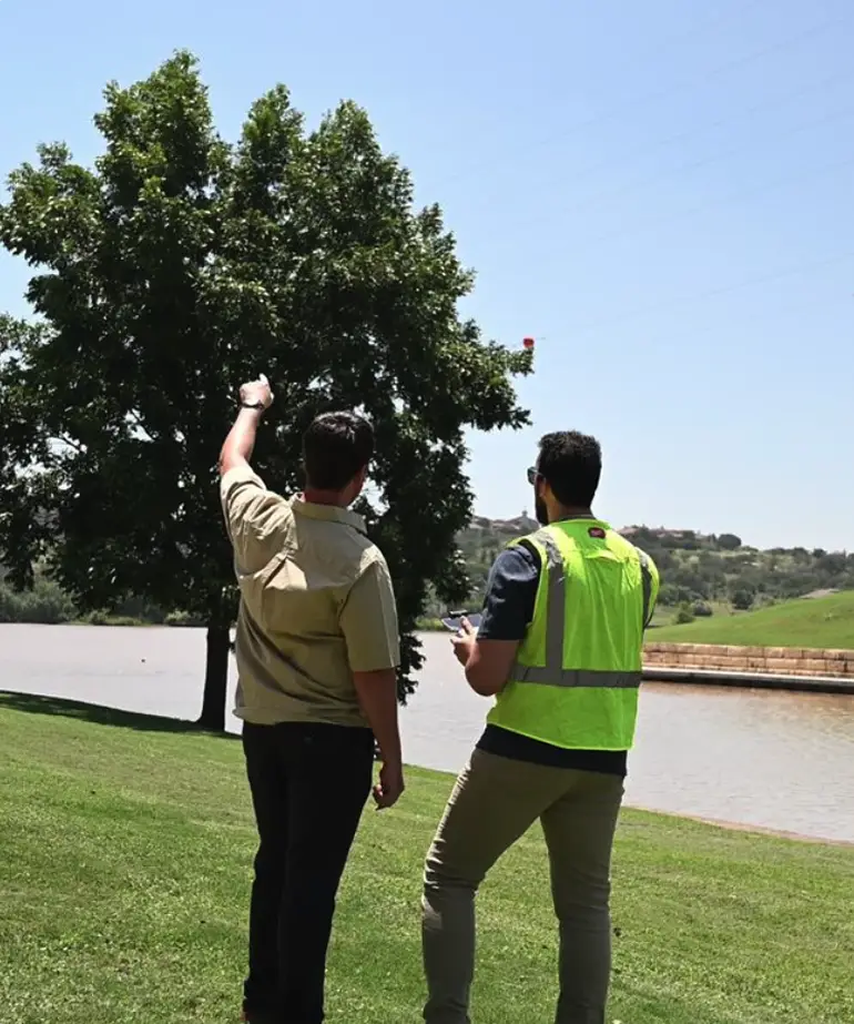 Two field engineers surveying a riverbank, one in a safety vest using a handheld device and the other pointing toward a tree.