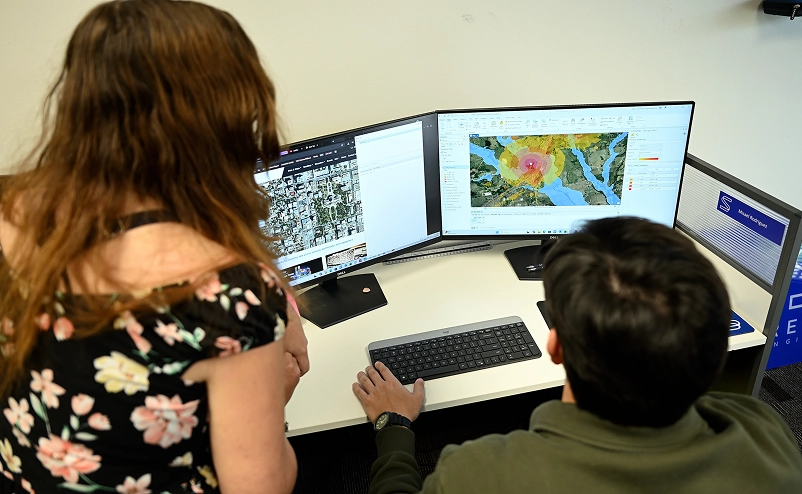 Engineers reviewing GIS flood mapping and satellite imagery on dual monitors in an office.